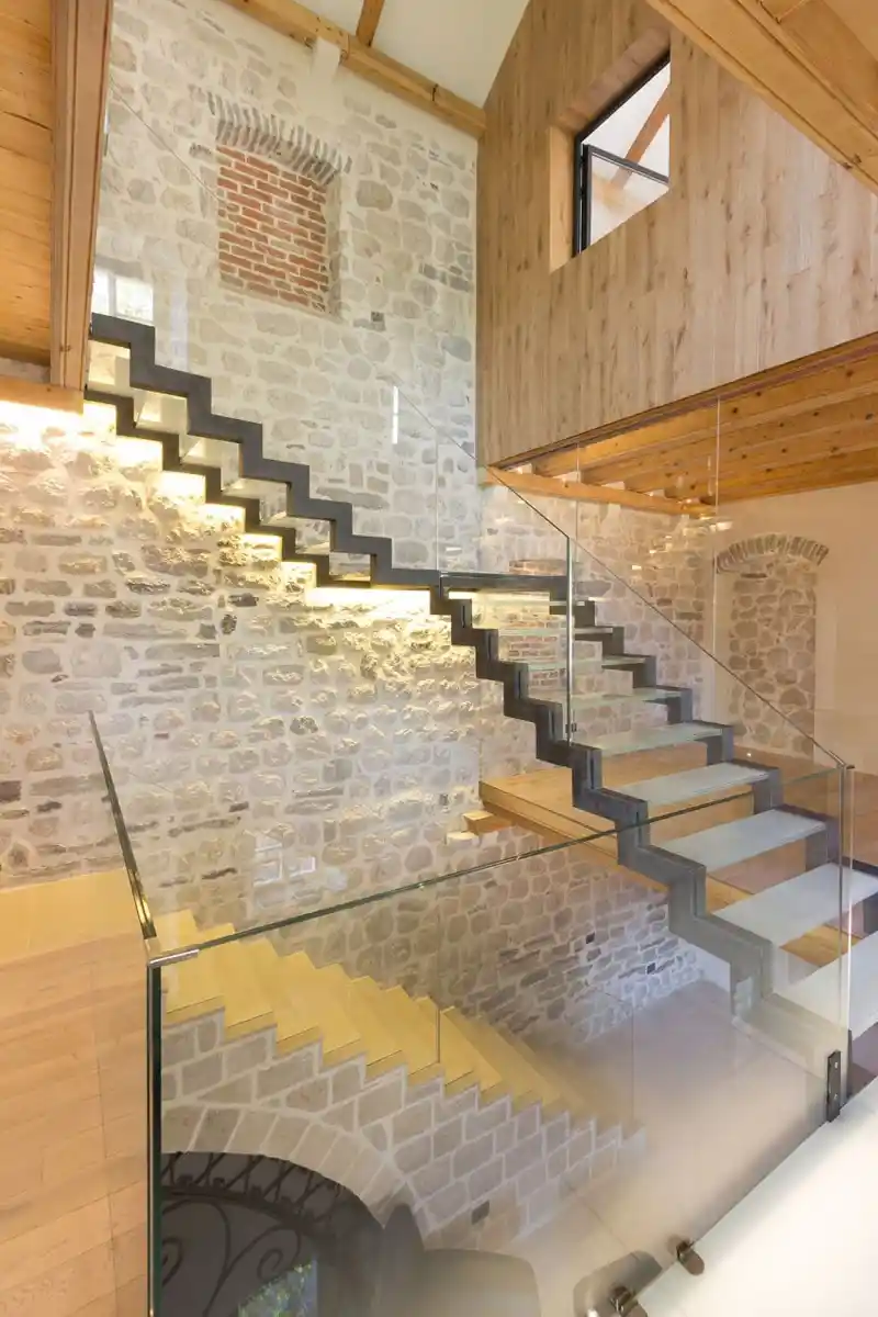 Escalier moderne en verre et bois avec mur en pierre reconstituée et plaquette de brique, illustrant notre expertise en matériaux nobles chez Orpa Matériaux (Ecotra Parement).